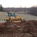 A yellow bulldozer excavating soil on a grassy field creates a long mound of earth. Leafless trees and rolling hills are visible in the background under a mostly cloudy sky, preparing the land for a future building. Kentucky Builders And Excavating