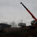 A large crane lifts a concrete structure over a wooden fence in a residential backyard, where building or excavating work is underway. A house sits on the left, and bare trees stand in the background beneath a cloudy sky. Kentucky Builders And Excavating