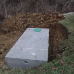 A partially buried concrete septic tank with a green circular lid sits in a dug-up area surrounded by dirt piles and grassy ground, near a wooded area, where excavating was done for a nearby building. Kentucky Builders And Excavating