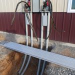 Two electrical panels mounted on a building exterior with conduit pipes running underground; several wires are exposed, and a corrugated metal sheet covers the trench below the panels from recent excavating. Kentucky Builders And Excavating