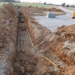 A long trench is dug in dirt for excavating utility cables or pipes. Nearby, a green utility box, yellow warning tape, and construction equipment sit on gravel next to a grassy field and a pole barn building. Kentucky Builders And Excavating