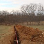A narrow trench dug through a grassy field for excavating a red pipe or conduit is partially buried inside. Piles of earth line both sides, while leafless trees and a cloudy sky set the scene—possibly groundwork for a future pole barn. Kentucky Builders And Excavating
