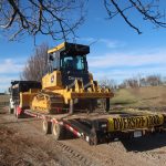 A yellow bulldozer used for excavating is loaded onto a flatbed trailer labeled OVERSIZE LOAD, being transported by a white truck on a rural dirt road with fields and bare trees in the background. Kentucky Builders And Excavating