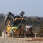 Two construction workers bend over while working near a small yellow excavator, excavating on a gravel surface at an outdoor pole barn building site, with trees and hills in the background. Kentucky Builders And Excavating