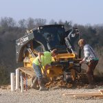 Two workers stand in front of a John Deere construction vehicle on a gravel site, excavating soil with shovels. Nearby are pipes and wooden boards for the building project, with trees visible in the background. Kentucky Builders And Excavating