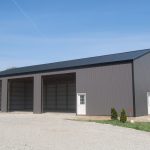 Large gray metal pole barn with three open garage bays and two white entry doors, set on a gravel lot with grass and small shrubs nearby, under a clear blue sky. Ideal for excavating or storage needs. Kentucky Builders And Excavating