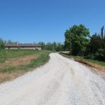 A gravel road curves through grassy land toward a single-story house and a nearby pole barn, with trees and a clear blue sky in the background. Kentucky Builders And Excavating