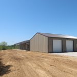 A spacious pole barn with two garage doors stands on a dirt lot under a clear blue sky, surrounded by trees and open space. Kentucky Builders And Excavating