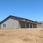 A large, light brown pole barn with several windows and a covered porch stands on a bare, dirt lot under a clear blue sky, with trees and a truck visible in the background. Kentucky Builders And Excavating