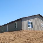 A tan metal pole barn with multiple windows and a white door stands on a bare dirt hill, showing signs of recent excavating under a clear blue sky. Kentucky Builders And Excavating