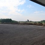 A flat, gravel-covered area bordered by piles of dirt hints at recent excavating, with green fields and silos in the distance under a partly cloudy sky. A covered pole barn and hanging flowers are visible on the right. Kentucky Builders And Excavating
