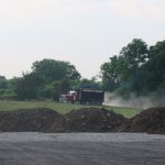 A red and black dump truck drives on a dirt road, raising dust near green trees and several piles of dirt, as part of an excavating project for a building. The sky above is partly cloudy. Kentucky Builders And Excavating