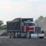 A red and black dump truck loaded with dirt is parked on a gravel area at a construction site, where excavating is underway for a new pole barn. Trees and a dirt road can be seen in the background. Kentucky Builders And Excavating