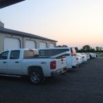 A row of white vehicles, including a pickup truck and several cars, is parked outside a large pole barn at sunset, with a grassy field in the background. Kentucky Builders And Excavating