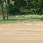A patch of ground with visible tire tracks in the dirt, possibly from recent excavating or building work, bordered by green grass, trees, and a line of rocks in front of a wooded area. Kentucky Builders And Excavating