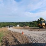 A gravel driveway under construction curves through a grassy area with dirt piles, as excavating work takes place. A bulldozer and small trailer are on site; an orange cone marks the edge of the gravel. Trees and blue sky frame the future pole barn location. Kentucky Builders And Excavating
