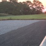 A gravel and asphalt parking area next to a concrete sidewalk, with a grassy field, mature trees, and the sun setting on the horizon—an ideal spot for building a pole barn after some excavating. Kentucky Builders And Excavating