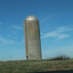 A tall, cylindrical concrete silo with a domed top stands on a grassy field, showing significant cracks and damage at the base. Nearby, a yellow bulldozer used for excavating and a pile of rebar sit under a blue sky with scattered clouds. Kentucky Builders And Excavating