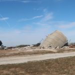 A collapsed grain silo lies in pieces on a dry, grassy field beside a dirt road, near an old pole barn. Debris and rubble are scattered around the fallen structure under a blue sky with wispy clouds. Kentucky Builders And Excavating