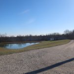 A gravel road curves toward a house and a small pond, with a pole barn nearby, all surrounded by grass and bare trees under a clear blue sky on a sunny day. Kentucky Builders And Excavating