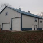 A large gray metal pole barn with blue trim, featuring three big garage doors, a small door, and an exterior staircase to a second-floor entrance. The building sits on grassy land with scattered leaves. Perfect for excavating equipment storage. Kentucky Builders And Excavating