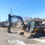 Two John Deere construction vehicles, a backhoe and a bulldozer, are parked on a dirt area beside a road under clear blue sky, ready for excavating work. Utility poles and some smoke are visible in the background. Kentucky Builders And Excavating