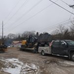 A black pickup truck with a flatbed trailer hauls excavating equipment, including a yellow excavator, near a pole barn on a cloudy day. A bulldozer operates nearby, snow covers the ground, and power lines run overhead. Kentucky Builders And Excavating