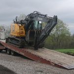 A yellow and black John Deere excavator is being loaded onto a flatbed trailer attached to a semi-truck on a gravel road, with grass, trees, and a pole barn building in the background under a cloudy sky. Kentucky Builders And Excavating