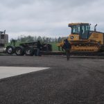 A red semi-truck with a flatbed trailer is parked on gravel, carrying a large yellow bulldozer—perfect for excavating. Two people are near the trailer, one working and one walking toward the truck beside a pole barn under an overcast sky. Kentucky Builders And Excavating