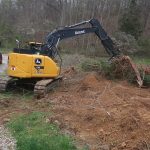 A yellow excavator is excavating tree roots and soil near a wooded area, with a dump truck nearby on a gravel path. The scene is outdoors with trees and grass in the background, preparing the site for building a pole barn. Kentucky Builders And Excavating