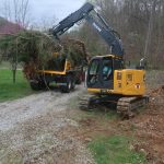 A yellow excavator loads a large bundle of tree branches and debris into a dump truck on a gravel driveway near a pole barn, surrounded by grass and trees in a rural area. Kentucky Builders And Excavating