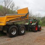 A green tractor with red wheels is attached to a large yellow dump trailer, tilted up on a gravel path beside trees and greenery. The setup looks ready for excavating work near a building or future pole barn on a cloudy day. Kentucky Builders And Excavating