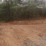 A cleared dirt area with visible tire tracks and a steep, freshly-cut embankment, likely prepared for building a pole barn, bordered by a forest of green trees. The ground appears recently excavated and the sky is overcast. Kentucky Builders And Excavating