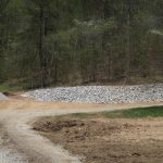 A winding gravel road curves through a clearing bordered by a pile of gray rocks and gravel, hinting at recent excavating, with dense, green-leaved trees rising on the sloped hillside. Kentucky Builders And Excavating