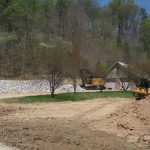 A construction site with two yellow excavators actively excavating piles of dirt and rocks, a house and pole barn in the background, all surrounded by trees on a sunny day. Kentucky Builders And Excavating