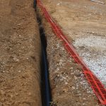 A trench dug in brown soil with a black pipe inside, partially covered by red warning mesh, showcases ongoing excavating work. A person stands in the background near rocks at the edge of the building site. Kentucky Builders And Excavating