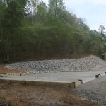 A concrete foundation slab for a pole barn is set on a gravel lot near a forested hillside, with piles of rocks behind it and a house with a garage in the background, suggesting recent excavating and ongoing building work. Kentucky Builders And Excavating