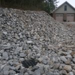 A large pile of gray rocks covers a sloped embankment beside a gravel driveway, with a metal drainage grate at the base. In the background, there is a house with a double garage and trees behind it, suggesting recent excavating or building work. Kentucky Builders And Excavating