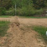 A narrow trench runs through a grassy yard toward a dirt driveway, likely for excavating work on a new building. Boxes of material are stacked on the right, with utility poles and wires visible among green trees in the background. Kentucky Builders And Excavating