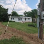 A white house is seen in the background with a trench excavating across the yard, leading to a utility pole with electrical boxes in the foreground. The sky is partly cloudy and trees surround the scene. Kentucky Builders And Excavating