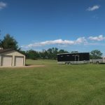 A black pickup truck with a large black trailer is parked on a grassy field near a beige pole barn building, under a blue sky with scattered clouds. Kentucky Builders And Excavating