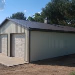 A beige metal pole barn building with two closed garage doors and a sloped dark roof sits on a concrete slab, surrounded by grass and trees under a blue sky. Kentucky Builders And Excavating