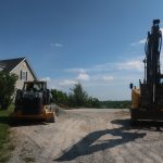 A yellow bulldozer and an excavator, likely used for excavating a new building or pole barn, are parked on a gravel driveway near a house, with trees and blue sky in the background. Kentucky Builders And Excavating