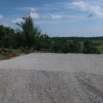 A gravel driveway extends towards a scenic, green countryside under a blue sky with scattered clouds, bordered by grass and trees on both sides—perfect access for future building projects or pole barn construction. Kentucky Builders And Excavating
