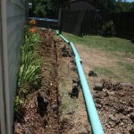 A large turquoise drainage pipe is being installed in a trench alongside a house, with dirt and rocks scattered around from the excavating. Orange flowers and green plants line the trench; a wooden fence and shed are visible in the background. Kentucky Builders And Excavating