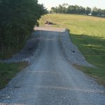 A gravel road slopes uphill through a grassy field with trees on the left. Rocks line both sides, and farm equipment is visible near the top of the hill, where a pole barn stands under a clear sky. Kentucky Builders And Excavating
