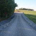 A gravel road lined with large stones runs through a grassy field, bordered by trees on the left and open land on the right, passing near a pole barn under a clear sky. Kentucky Builders And Excavating