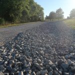 A gravel road curves to the left, bordered by larger gray rocks on one side. Trees and grass line the road under a clear, sunny sky, leading toward a pole barn in the distance. Kentucky Builders And Excavating