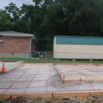 A concrete foundation with red rebar grid is being laid for a building project, bordered by wooden forms. A brick house, a green-roofed shed, and trees are in the background. An orange traffic cone is near the edge. Kentucky Builders And Excavating