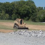 A bulldozer with tan paint is excavating dirt on a construction site for a pole barn, surrounded by grass, gravel, piles of rocks, and trees beneath a clear blue sky. Kentucky Builders And Excavating